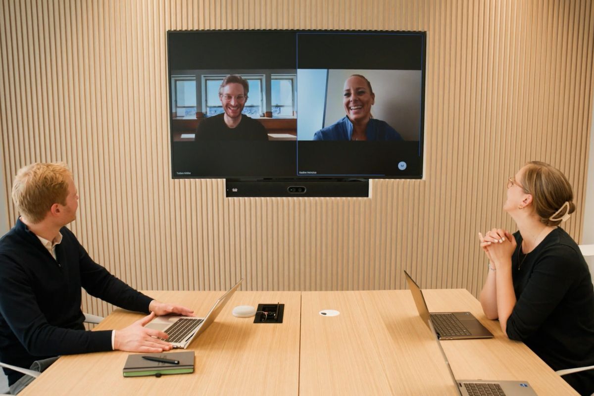 Hybrides Arbeiten: Zwei Personen sitzen im Raum, auf dem Bildschirm an der Wand sind zwei weitere Personen zu sehen
