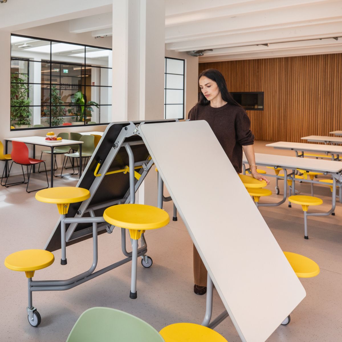 Frau klappt großen Tisch mit Sitzhockern zusammen, in der Cafeteria der Frankfurt International School, geplant und eingerichtet von designfunktion